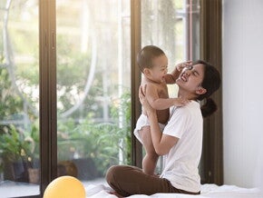 Smiling Asian mom holding her clapping baby while seated on cushions on the floor
