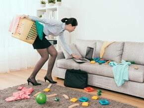 Young Asian mom stressed at messy living room with toys and laundry, trying to tidy up.