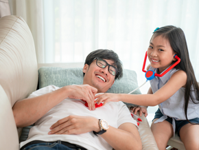 Young Asian girl listens to her father's heartbeat with a toy stethoscope, pretending to do dad’s medical checkups