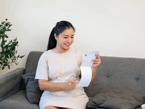 Young Asian pregnant woman on the sofa, holding her tummy and looking at her ultrasound scan at 28 weeks pregnant