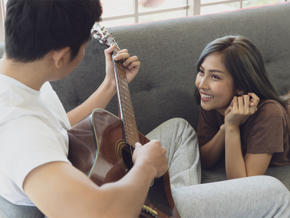Young Asian woman and man sitting on the couch while man plays guitar