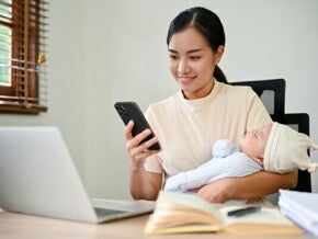 Young freelancing Asian mom checks PhilHealth requirements on her mobile with her baby in her arms.