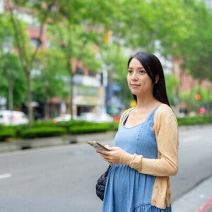A 17-week-pregnant Asian woman holding her smartphone while standing on the street.