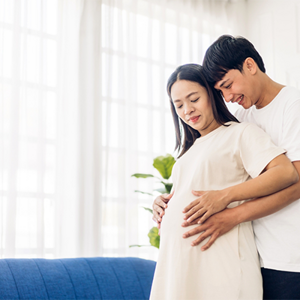 A 26 weeks pregnant woman holding her belly while her husband hugs her from behind in their home