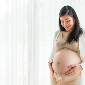 A 31 weeks pregnant Asian woman standing by the window, holding her large belly bump