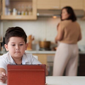 A little Asian boy watching a show on a tablet while his mother is cooking dinner in the background A little Asian boy watching a show on a tablet while his mother is cooking dinner in the background