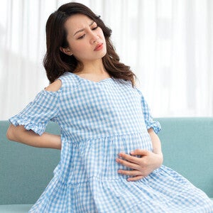A pregnant Asian woman is sitting on the sofa and holding her belly in discomfort due to cramps during pregnancy