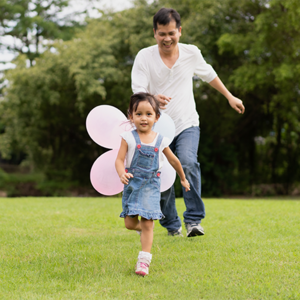 A smiling Asian preschooler girl running in the park with her dad for a budget-friendly day out. 
