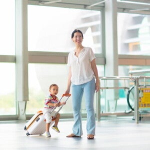A young Asian mom with her young toddler riding on her luggage at the airport.