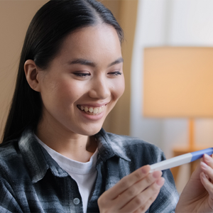 A young Asian woman smiling as she looks at a pregnancy test and curious about the stages of pregnancy.