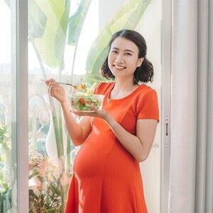 A young Asian woman standing by the window and holding a bowl of salad as part of her healthy pregnancy meal plan A young Asian woman standing by the window and holding a bowl of salad as part of her healthy pregnancy meal plan