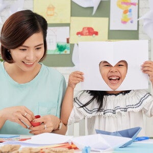 A young girl showing a heart cutout on paper, doing arts and craft for kids at home with her mom.