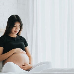 A young pregnant Asian woman holding her belly and seated in bed, alert to the signs of labor A young pregnant Asian woman holding her belly and seated in bed, alert to the signs of labor