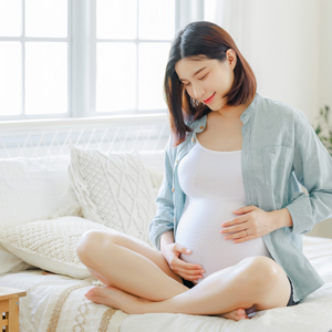 A young, pregnant Asian woman sitting cross-legged on the bed, touching her belly, aware of the importance of recognizing preeclampsia symptoms A young, pregnant Asian woman sitting cross-legged on the bed, touching her belly, aware of the importance of recognizing preeclampsia symptoms