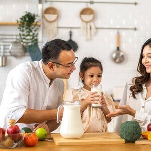 Asian couple watching their little girl drink milk from a glass in the kitchen.