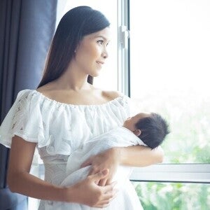 A young Asian mom holding her baby and looking out the window to see if it’s safe to open for a healthy environment.