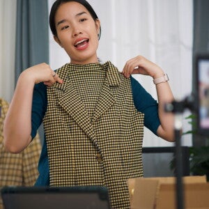 A young Asian woman sitting at a kitchen table with papers, a laptop, and a phone, looking happy. 