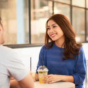 A young Asian single mom seated on a café table and smiling at her date while holding her juice.