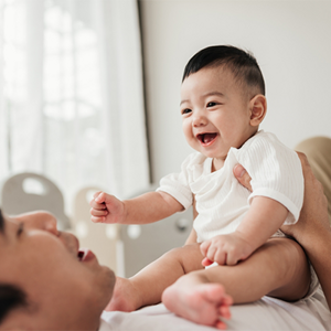 Asian dad lying on bed talks to his smiling baby who's sitting on his stomach