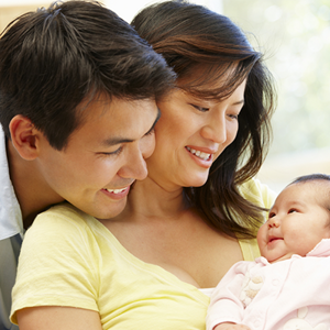 Asian father embraces his wife as she carries their infant who smiles at them, a new step in the child development stages.