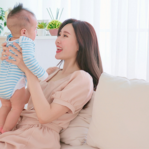 Asian mother holds and talks to her baby on the sofa at home, feeling healthy with proper postpartum care.