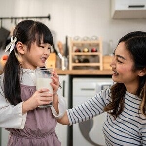 Asian preschooler girl with a glass of milk as her mom smiles at her in the kitchen, happy she’s getting protein in milk