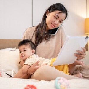 Asian working mom with her baby in bed talks on the phone while looking at documents.