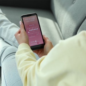 Close-up photo of a woman seated on the couch and using a period calculator on her phone.