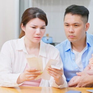 A worried Filipino couple sitting at a table with their baby and their bills, discussing finances and their source of income.