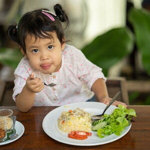 Cute Asian little girl eats fried rice with tomatoes and green leafy vegetables on the side Cute Asian little girl eats fried rice with tomatoes and green leafy vegetables on the side