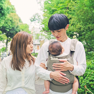Young Asian dad with wife beside him brings his little one outdoors in a soft-structured baby carrier. Young Asian dad with wife beside him brings his little one outdoors in a soft-structured baby carrier.