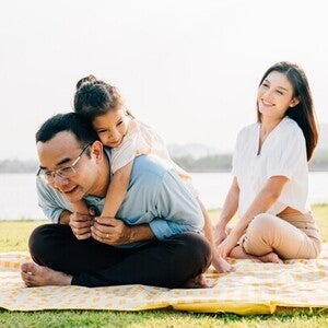 Little Asian girl hugs her dad as they spend an afternoon in the park for Father’s Day, while mom smiles and watches.