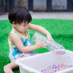 Little Asian boy playing with a DIY Montessori toy, like an empty plastic bottle and beads in a container