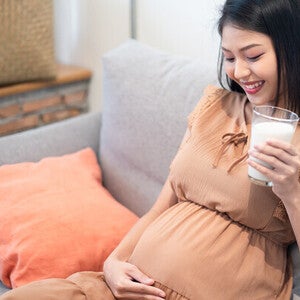 Pregnant Asian woman on the sofa, looking at and touching her belly while holding a glass of milk for pregnant women