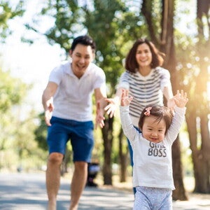 Cute Asian toddler wearing a “girl boss” shirt lifts her arms as her parents reach out to her in the background.