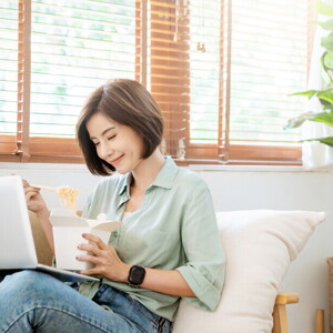Young Filipina experiences a food craving and enjoys takeout while sitting on the sofa with her laptop on her lap.