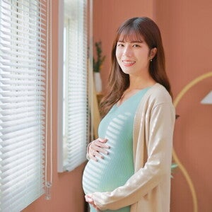 Asian woman, who is 39 weeks pregnant, holds her belly bump and smiles at the camera.