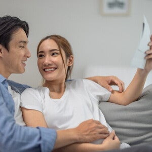 Asian woman at 18 weeks pregnant holds up up an ultrasound scan to show to her husband who’s beside her on the sofa. 