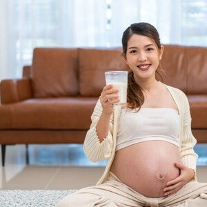 A Practical Guide to Calcium in Pregnancy Pregnant Asian woman holding a glass of milk in front of brown sofa.