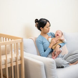 A young Asian mom carrying her little one while sitting on the sofa, wondering how to make a baby sleep.