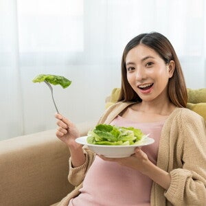 Asian pregnant woman eating greens on sofa.