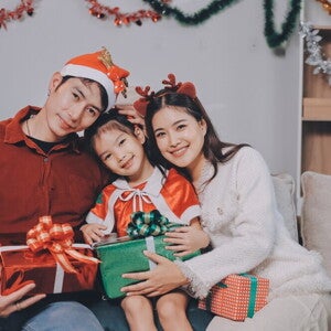 A young Asian couple with their little girl posing for a Christmas photoshoot while holding Christmas presents and wearing holiday headwear.