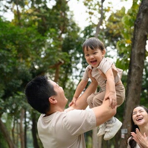 Cropped photo of a dad outdoors lifting his little boy in the air as a form of risky play that builds self-confidence in kids while mom looks on 