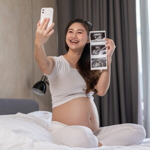 Pregnant Asian takes a selfie in bed while holding her pregnancy ultrasound picture.