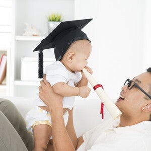 Asian dad smiles at his baby who has a graduation cap on as he bites on a diploma. 