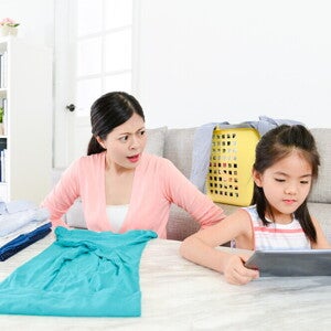 A young Asian mom is getting angry at her little girl playing on a tablet as she folds laundry.
