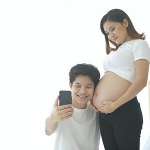 Young Asian pregnant couple taking selfie by a window for bright and beautiful baby bump pictures.