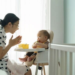 Young Asian mom claps her hands as she watches her baby eating in a high chair. Young Asian mom claps her hands as she watches her baby eating in a high chair.