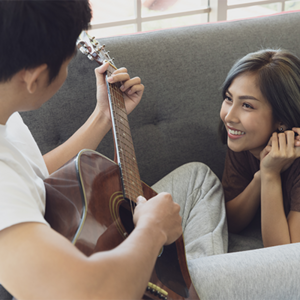 Young Asian woman and man sitting on the couch while man plays guitar