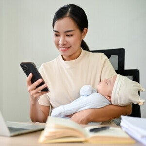 Young freelancing Asian mom checks PhilHealth requirements on her mobile with her baby in her arms.